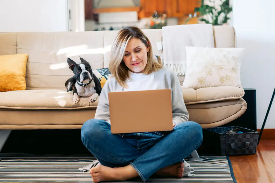 A woman sits on the floor with a laptop while a Boston terrier lies on the couch behind her.